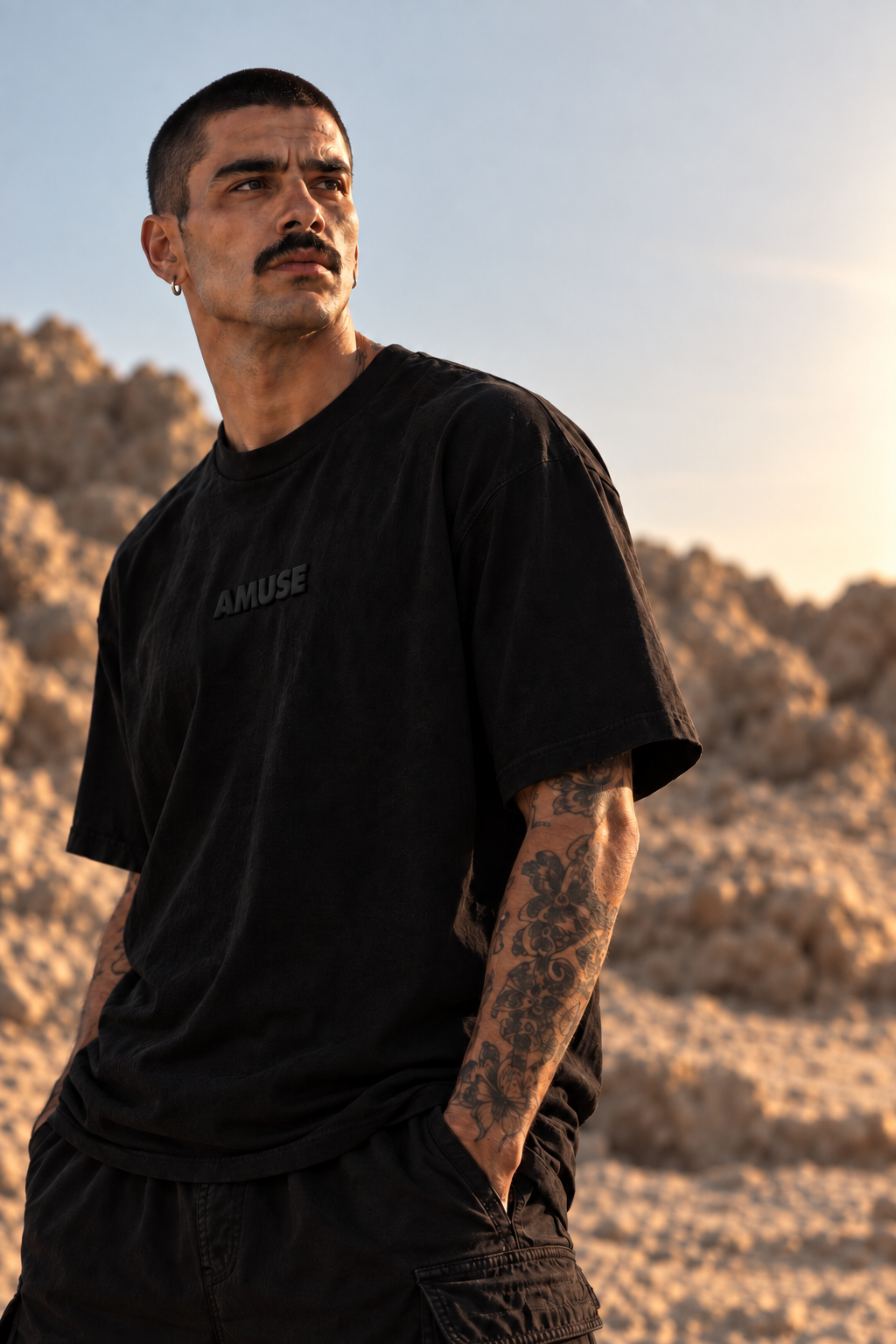 A man wearing a black LMNO t-shirt and black cargo pants, standing in a desert landscape during golden hour.