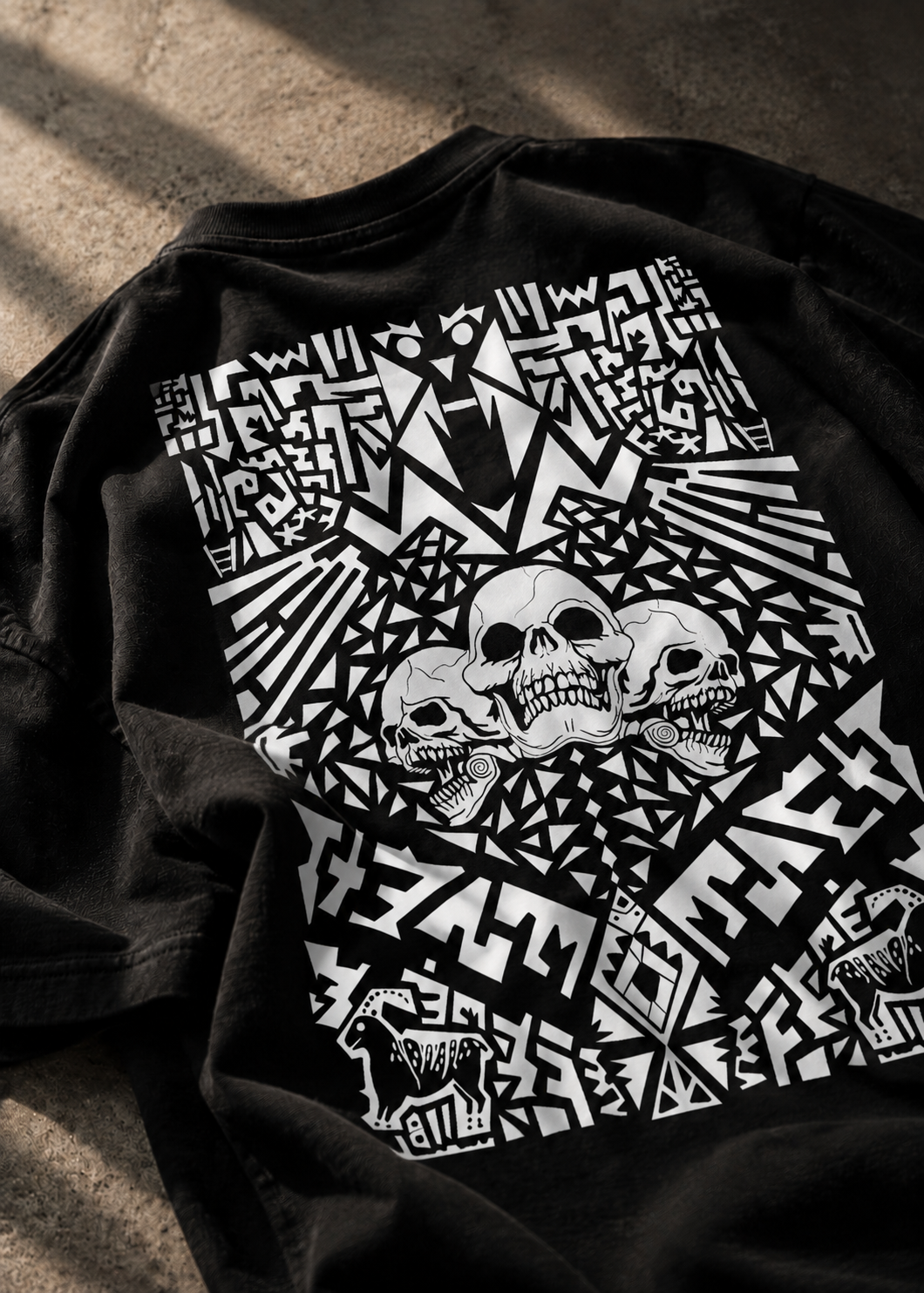 A black graphic LMNO t-shirt laid flat on a textured concrete surface, featuring a large white geometric print with three skulls.