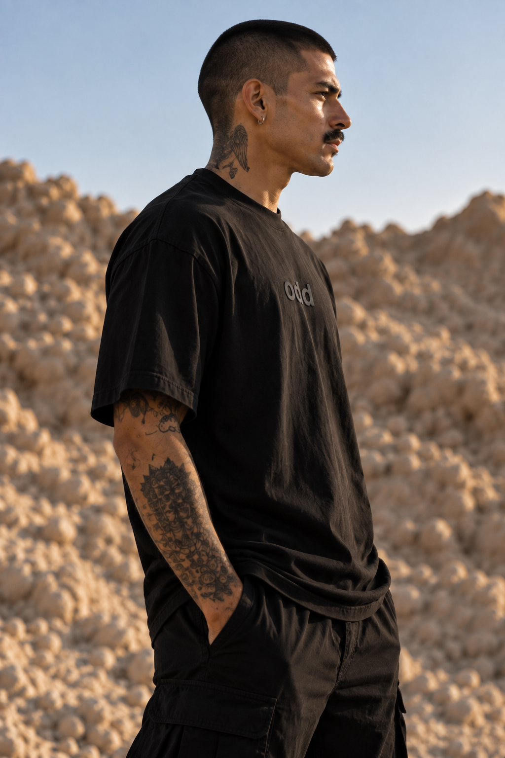 A man in a black LMNO t-shirt and black cargo pants standing in a rocky desert landscape under a clear blue sky.