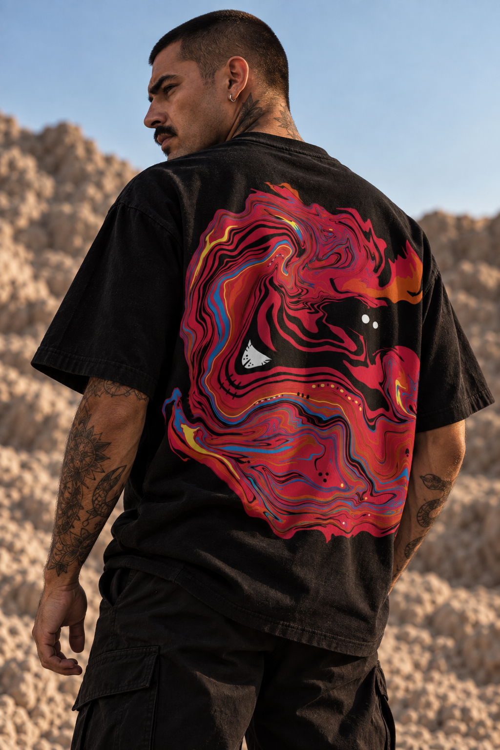 A man wearing a black LMNO t-shirt with a large, vibrant red, pink, and blue abstract fluid art graphic on the back, photographed outdoors against a rocky desert landscape.