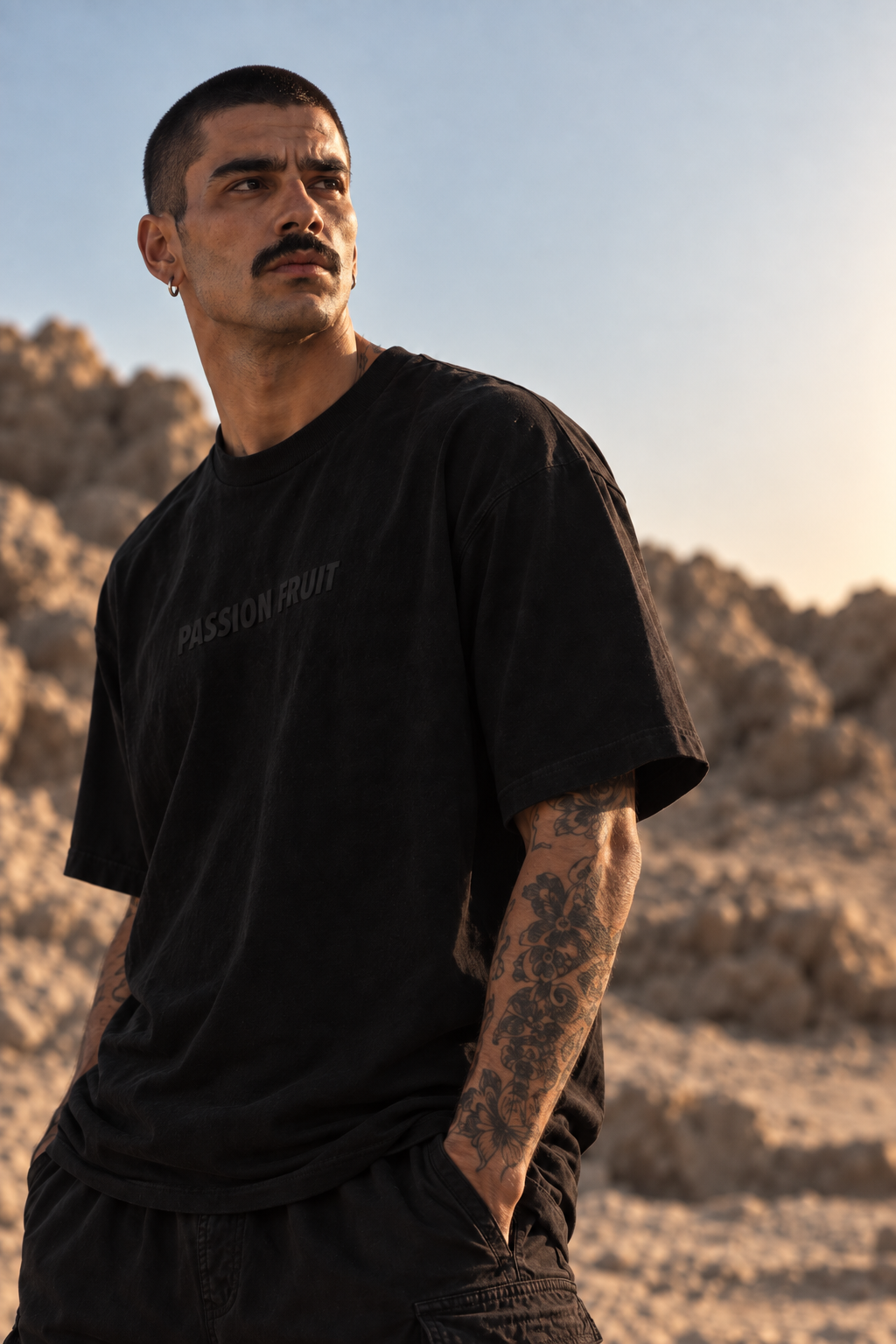 A man wearing a black LMNO t-shirt with 'PASSION FRUIT' printed on the chest, standing in a desert landscape at sunset.