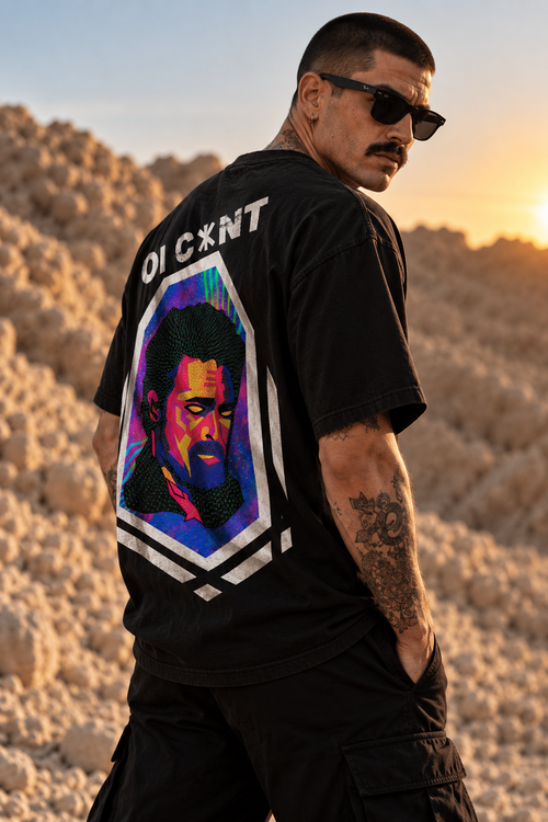 A man wearing a black LMNO t-shirt with a large multicolor portrait print on the back, standing in a rocky desert landscape at sunset.