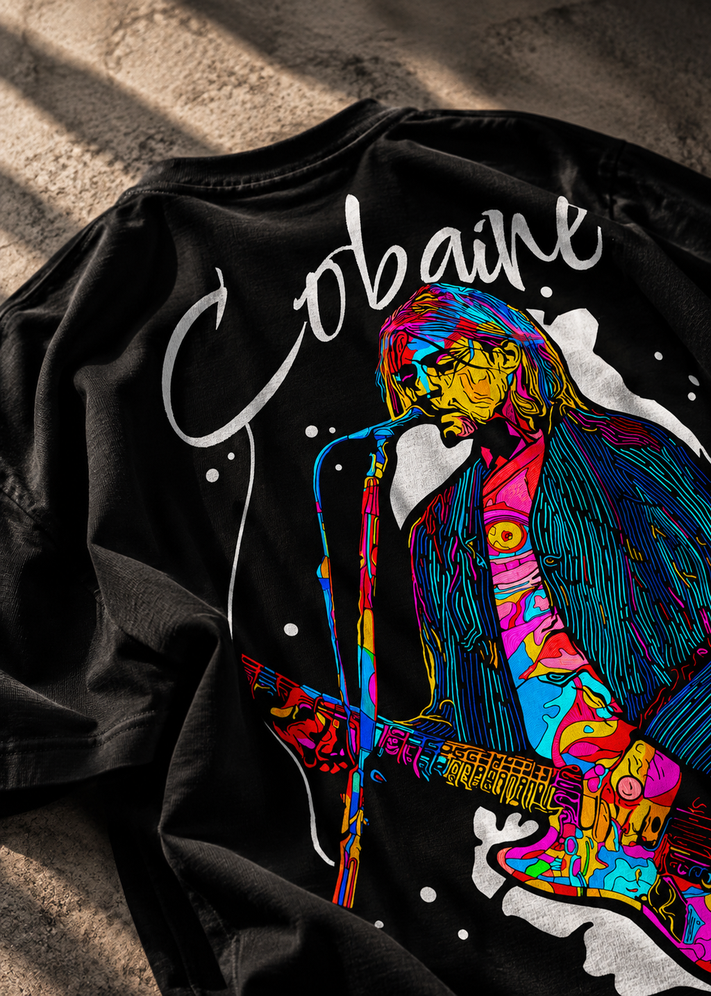 A black graphic LMNO t-shirt featuring a colorful, stylized illustration with the word 'Cobaine' in white script laid flat on a textured concrete surface.