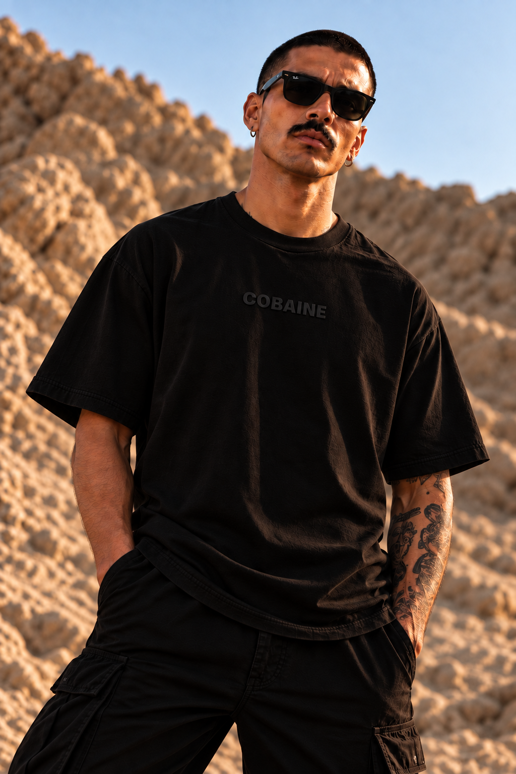 A man wearing a black LMNO t-shirt with the word 'COBAINE' printed in dark gray on the chest, styled with black cargo pants and sunglasses, standing outdoors against a rocky desert background.