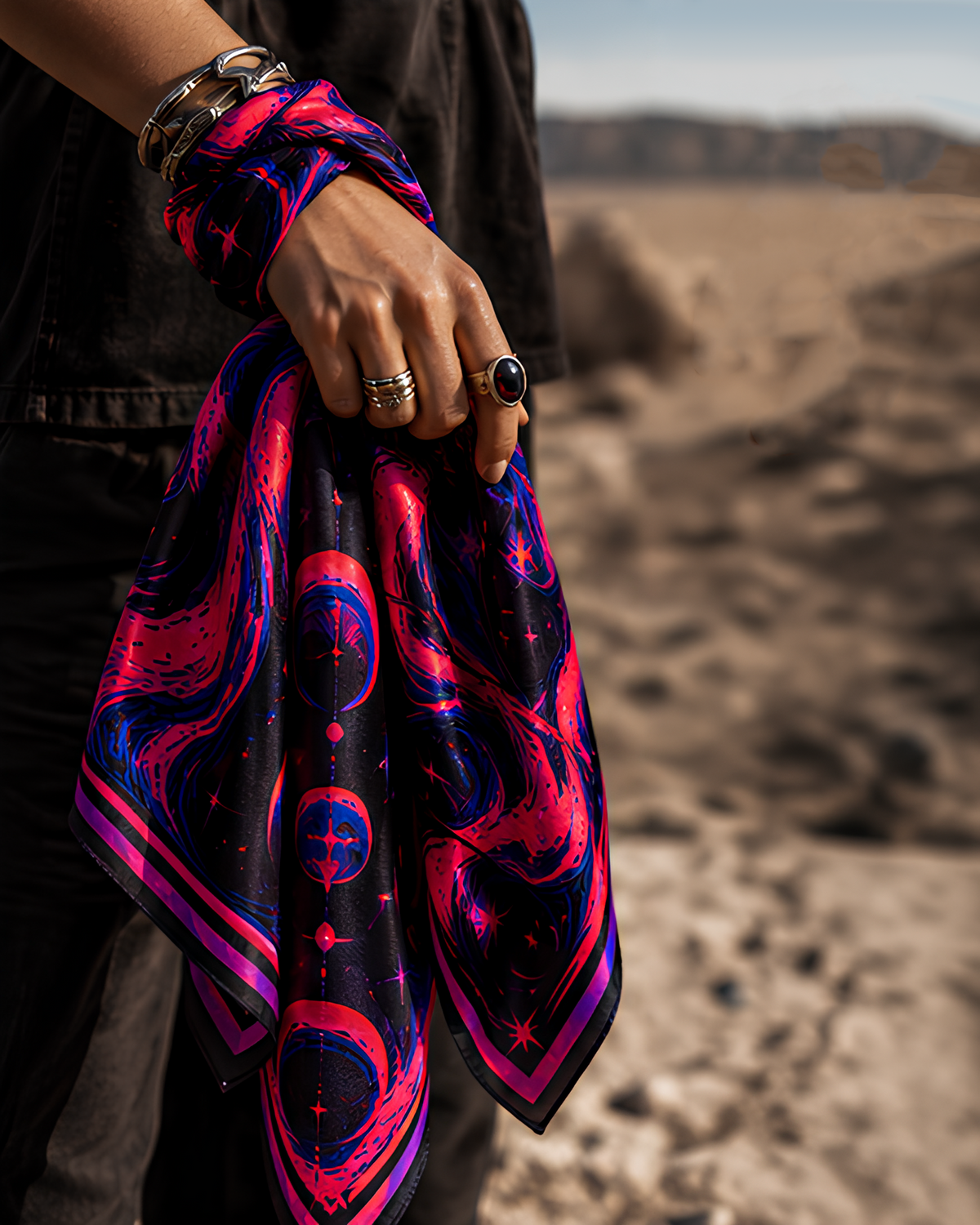 A person outdoors in a desert-like landscape holds up a square silk LMNO scarf with a cosmic print featuring pink, purple, and blue celestial motifs on a black background.