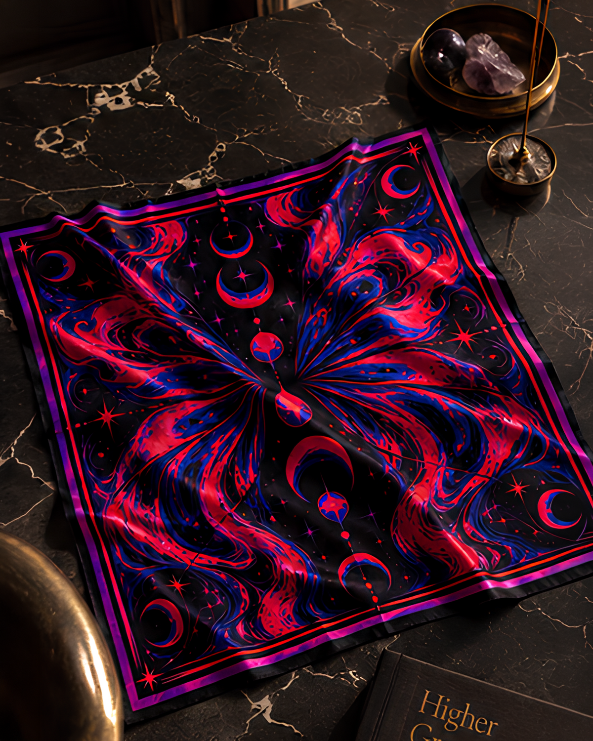 A square LMNO psychedelic scarf with a vibrant cosmic butterfly pattern in neon pink, red, and blue on a black background, draped on a black marble table with crystals and a brass incense holder nearby.