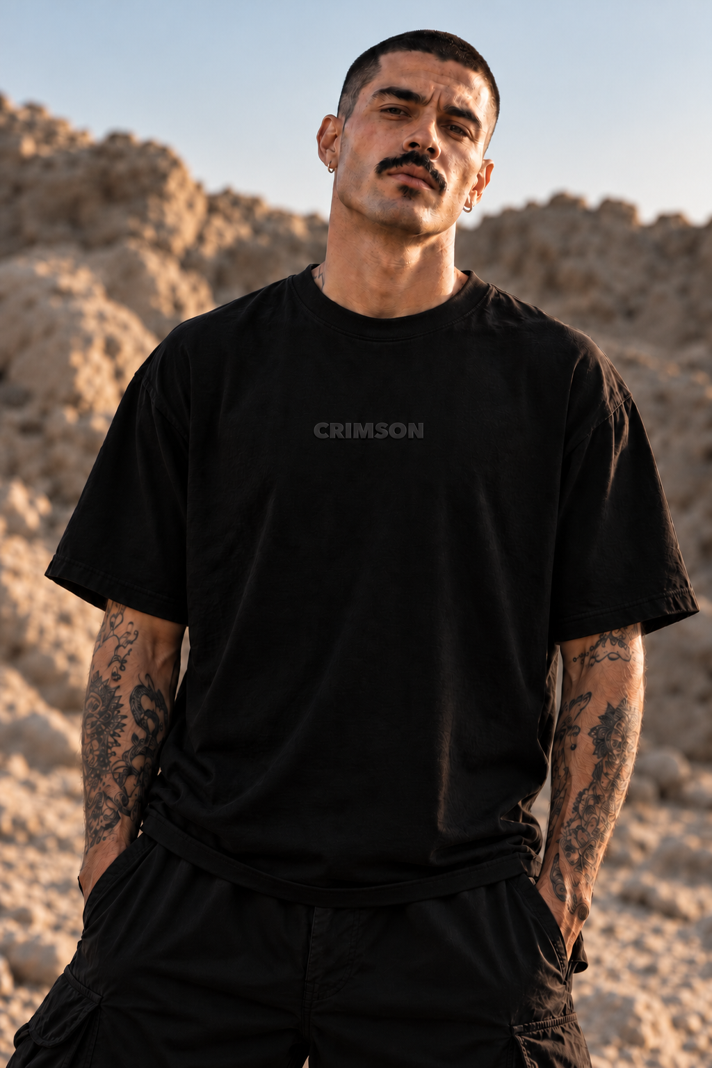 A man with a mustache and tattoos wearing a black LMNO t-shirt with the word 'CRIMSON' printed in gray on the chest, standing outdoors against a sandy, rocky background.