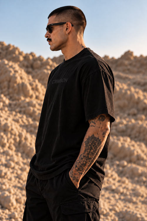 A man in a black LMNO t-shirt with 'CRIMSON' printed on the chest, standing in a desert landscape with rocky hills under a clear blue sky.