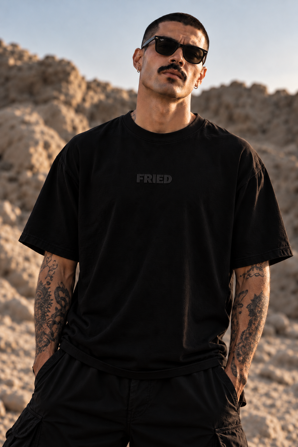 A man wearing a black LMNO t-shirt with a subtle dark gray 'FRIED' logo on the chest, styled with black cargo pants and sunglasses, standing in a sandy desert landscape.