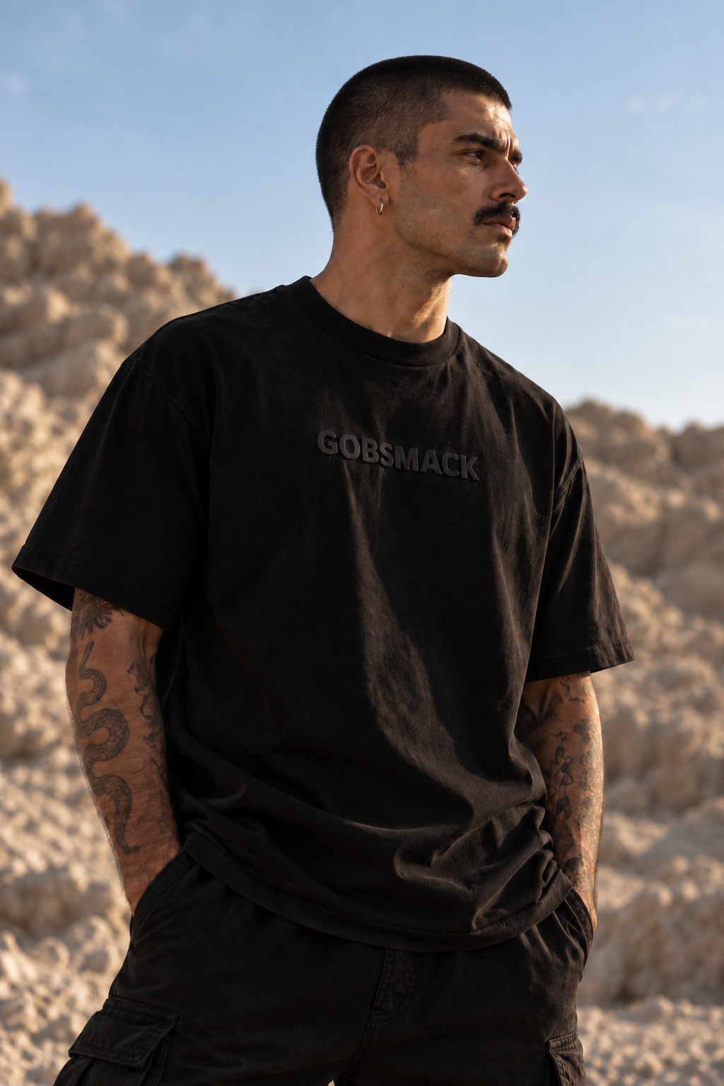 A man wearing a black LMNO t-shirt with a dark gray "GOBSMACK" text on the chest, standing outdoors in a rocky desert landscape under a clear blue sky.