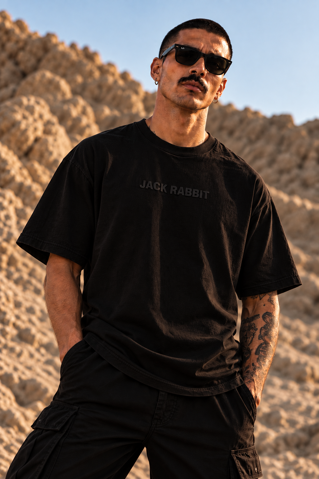 A man wearing a black LMNO t-shirt with 'JACK RABBIT' printed on the chest, posing outdoors against a rocky desert background.