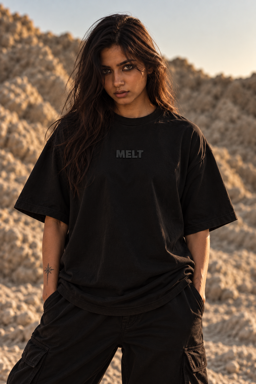 A woman wearing an oversized black LMNO t-shirt with the word 'MELT' printed in gray on the chest, standing in a sandy desert landscape during golden hour.