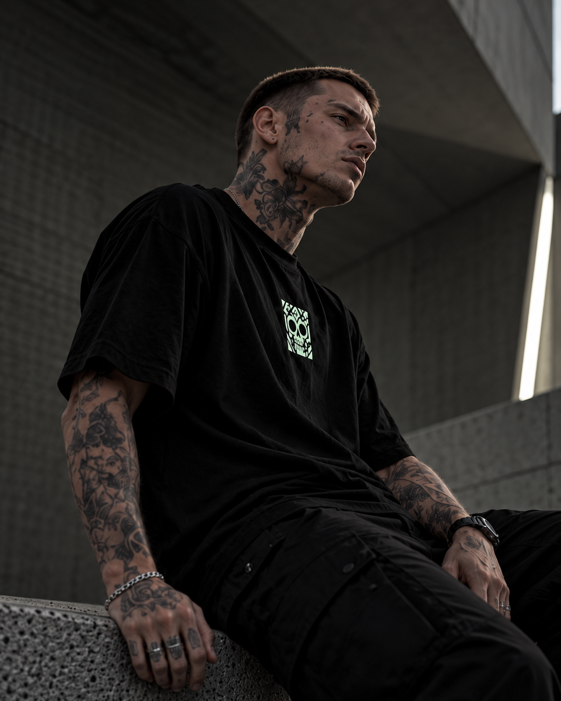 A tattooed man wearing a black oversized LMNO t-shirt with a small green skull graphic on the chest, sitting on a concrete ledge in a modern concrete structure.