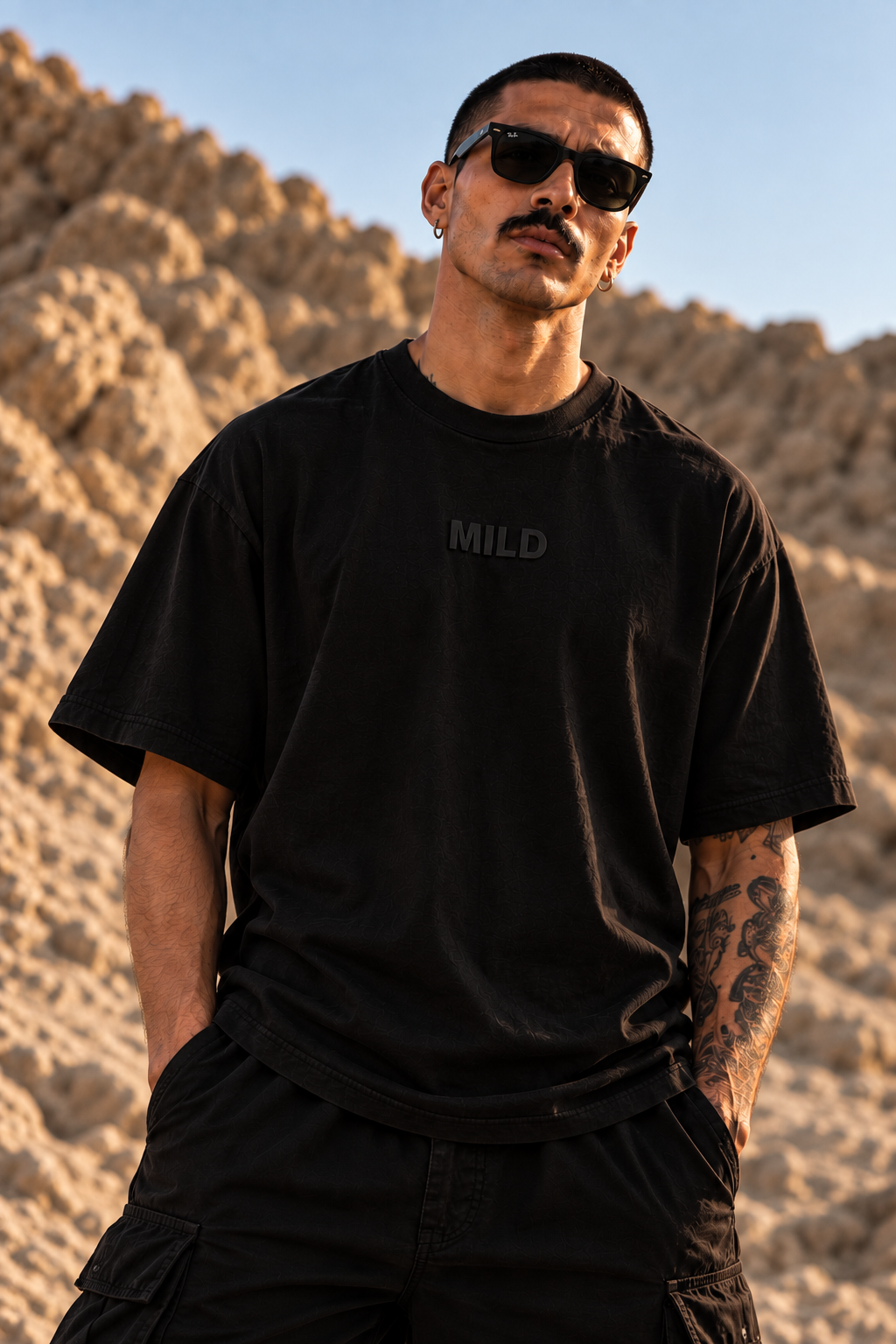 A man wearing a black oversized LMNO t-shirt with the word 'MILD' printed in dark gray on the chest, set against a rocky desert background.