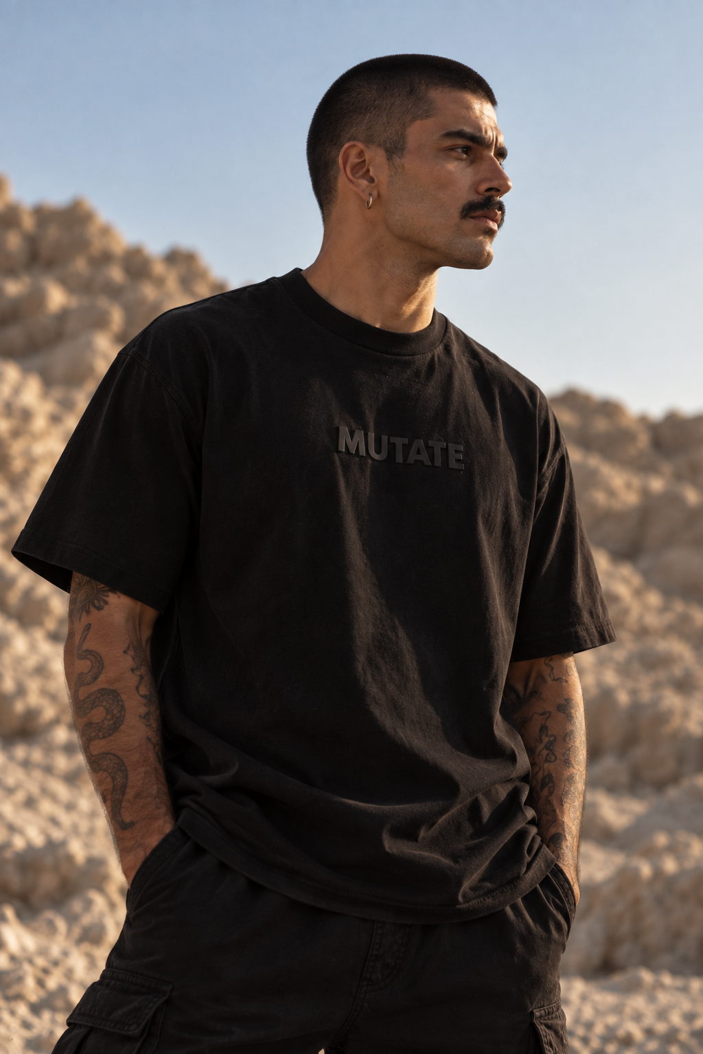 A man wearing a black LMNO t-shirt with the word 'MUTATE' printed in a dark gray on the chest, standing outdoors against a rocky desert background.