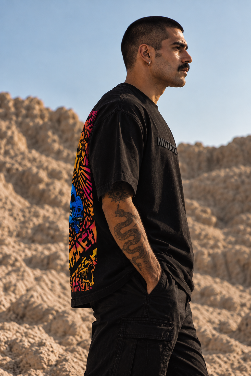 A man in a desert landscape wearing a black LMNO t-shirt with a colorful graphic print on the back and a small black logo on the chest, paired with black cargo pants.