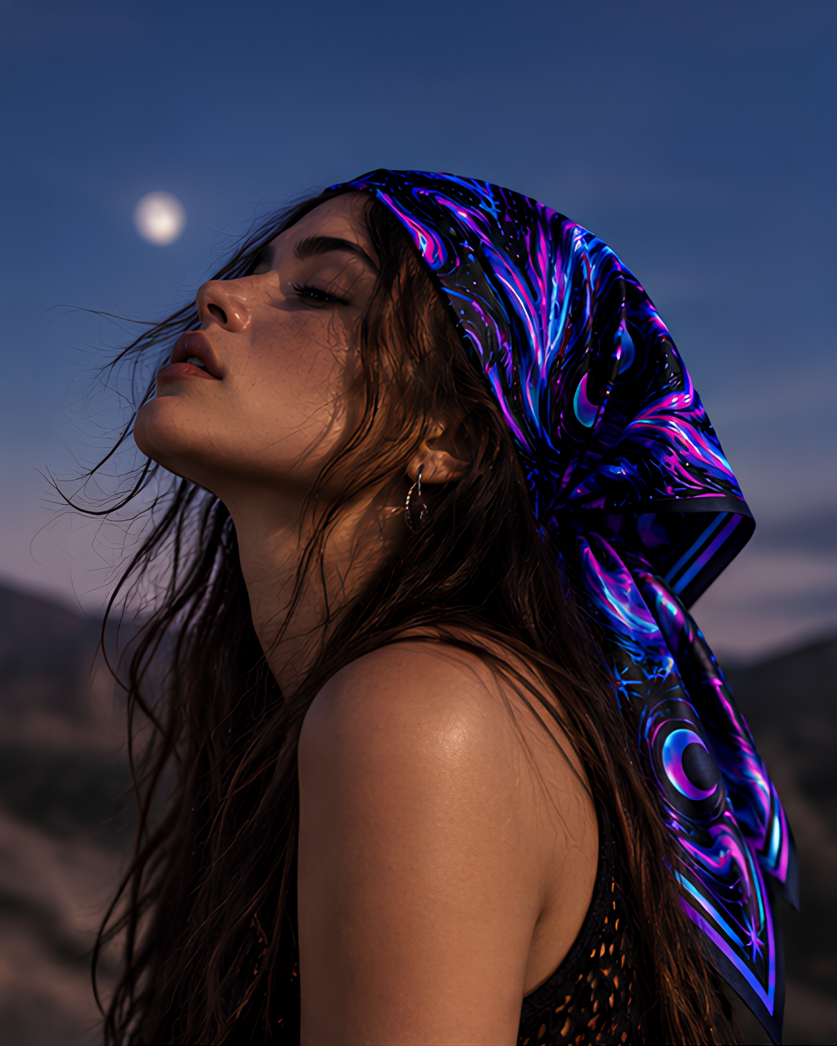 A woman outdoors at dusk wearing a vivid patterned LMNO headscarf in purple, blue, and black, with mountains and a moon visible in the background.