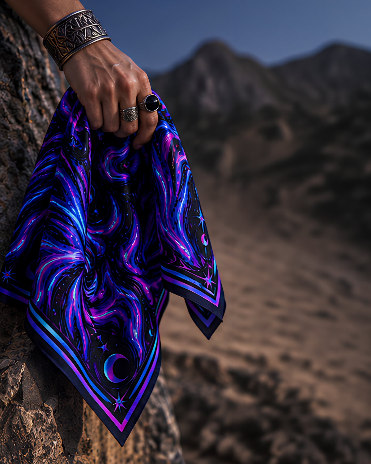 A hand wearing a silver cuff bracelet and rings holds a square LMNO scarf with a vivid cosmic galaxy print in purple, blue, and black, set against a blurred desert mountain landscape.