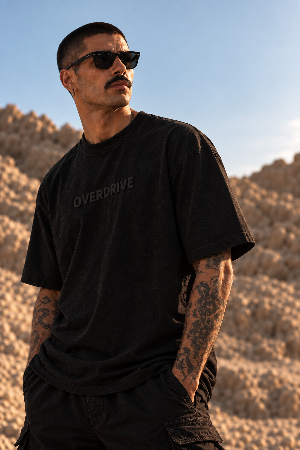 A man wearing a black graphic LMNO t-shirt with 'OVERDRIVE' printed on the chest, standing in a desert landscape at sunset.