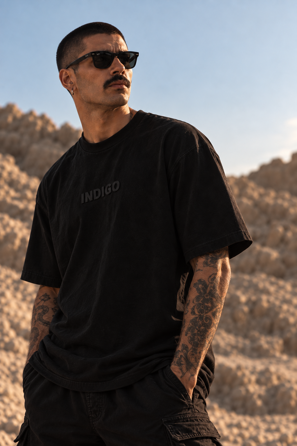 A man wearing a black LMNO t-shirt with 'INDIGO' printed on the chest, styled with black cargo pants and sunglasses, standing in a rocky desert landscape.