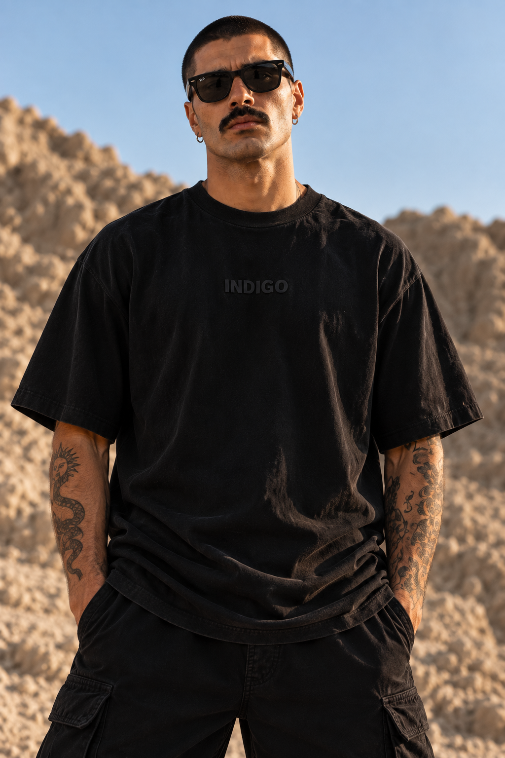 A man wearing a black LMNO t-shirt with a subtle 'INDIGO' logo on the chest, set against a desert landscape background.