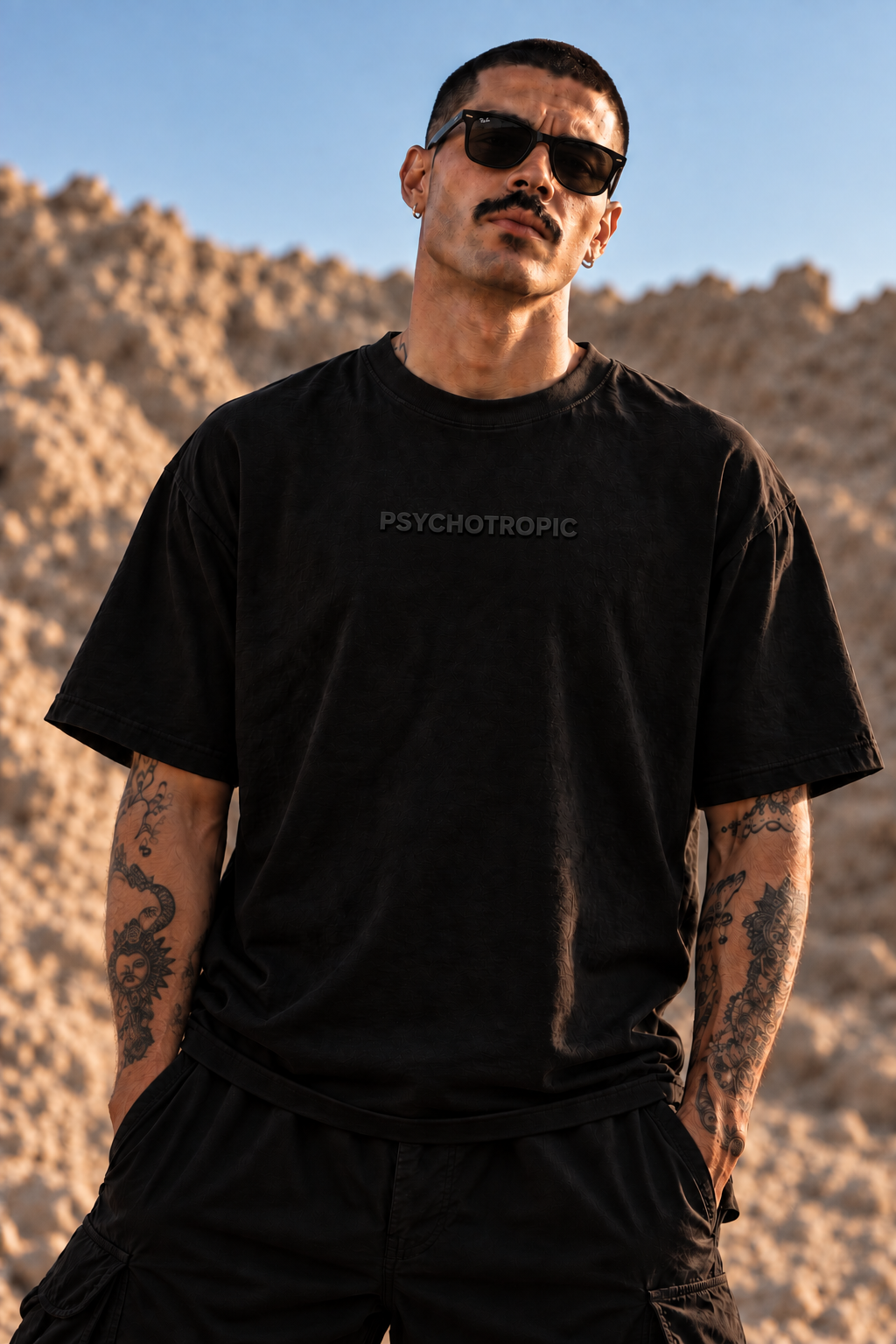 A man wearing a black LMNO t-shirt with the word 'PSYCHOTROPIC' printed in gray on the chest, set against a desert landscape background.