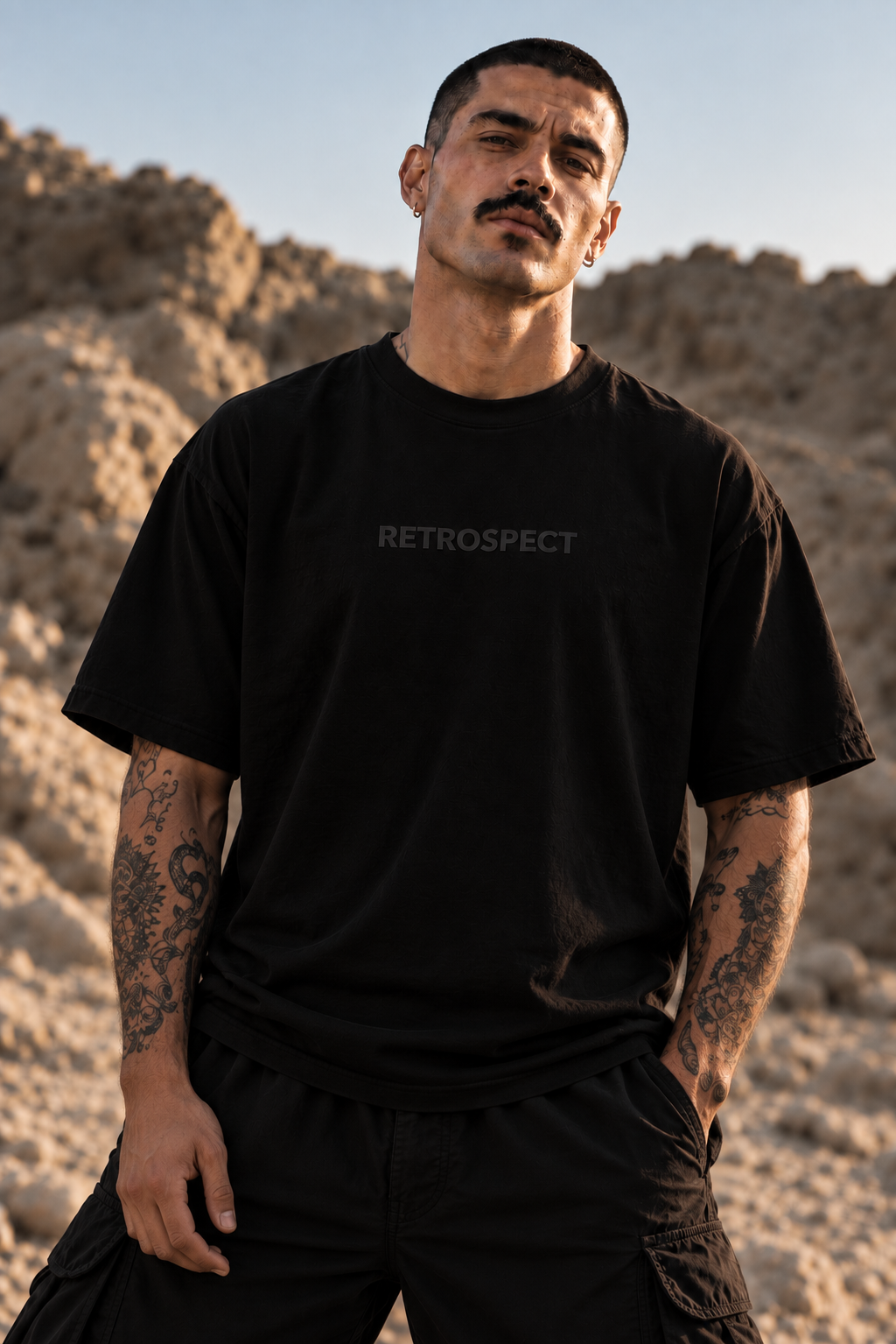 A man with a mustache wearing a LMNO black t-shirt with the word 'RETROSPECT' puff printed in gray on the chest, standing outdoors against a rocky desert background.