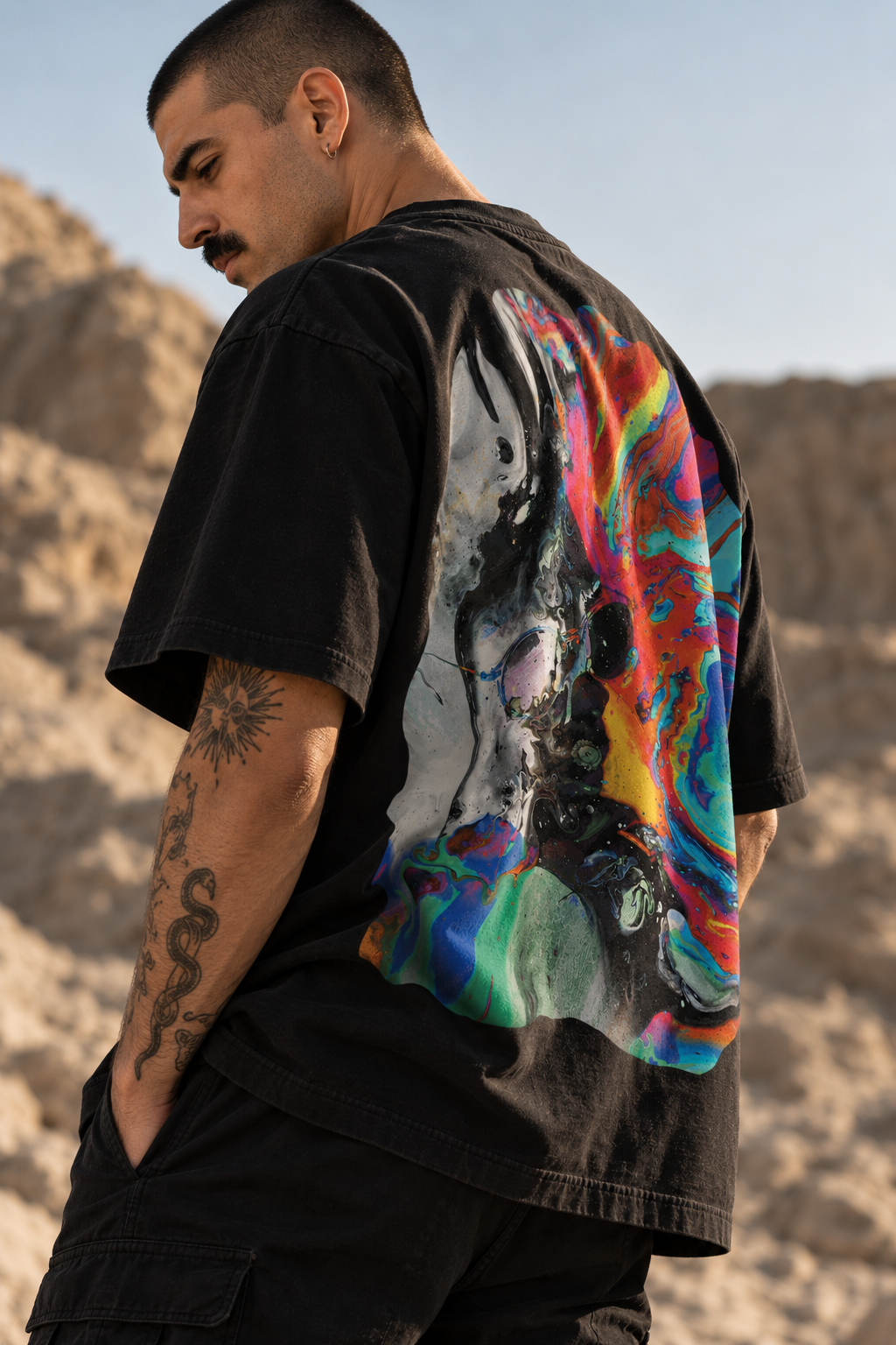A man wearing a LMNO black graphic t-shirt with an abstract print on the back, photographed outdoors against a sandy desert background.