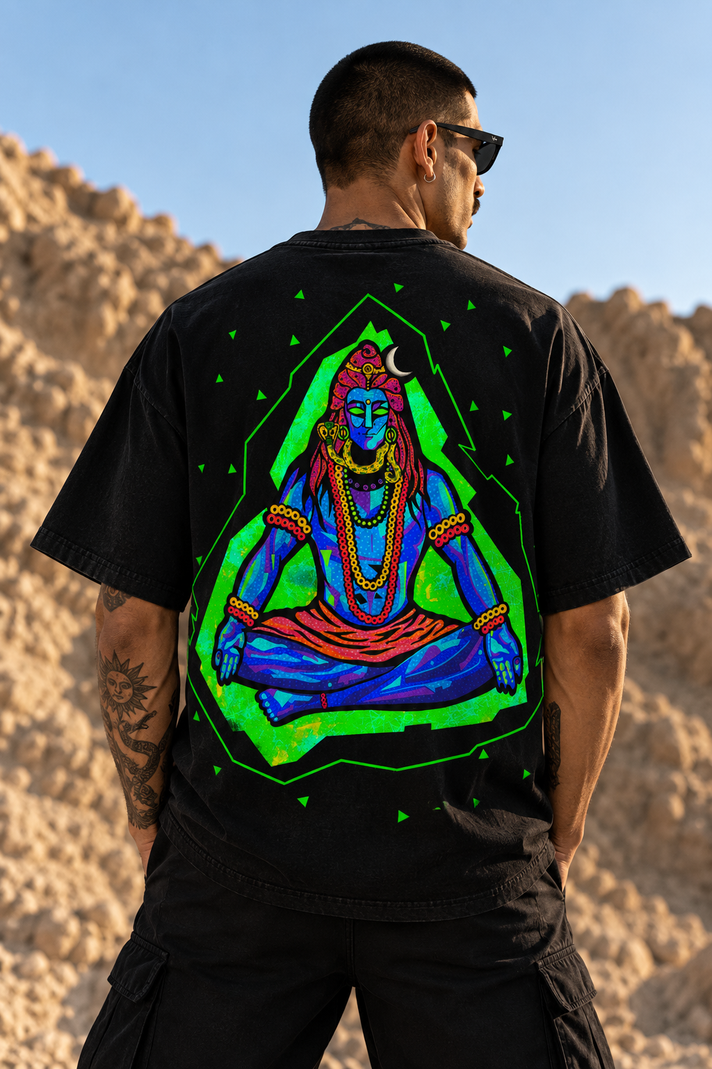 A man wearing a black LMNO t-shirt with a large, vibrant graphic of a meditating Lord Shiva on the back, set against a desert rock landscape.