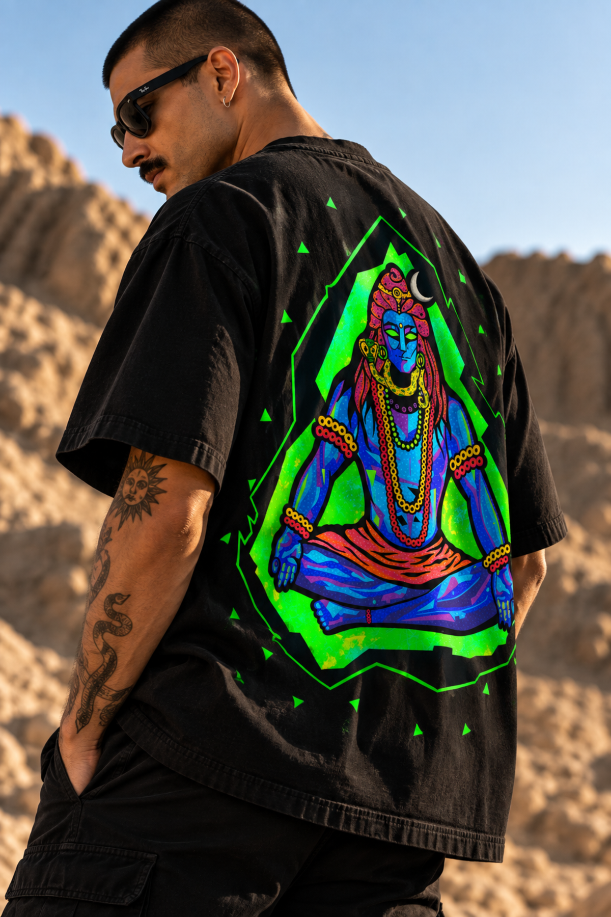 A man wearing a black LMNO t-shirt with a large, vibrant graphic of Lord Shiva meditating in neon green and blue, set against a desert landscape background.