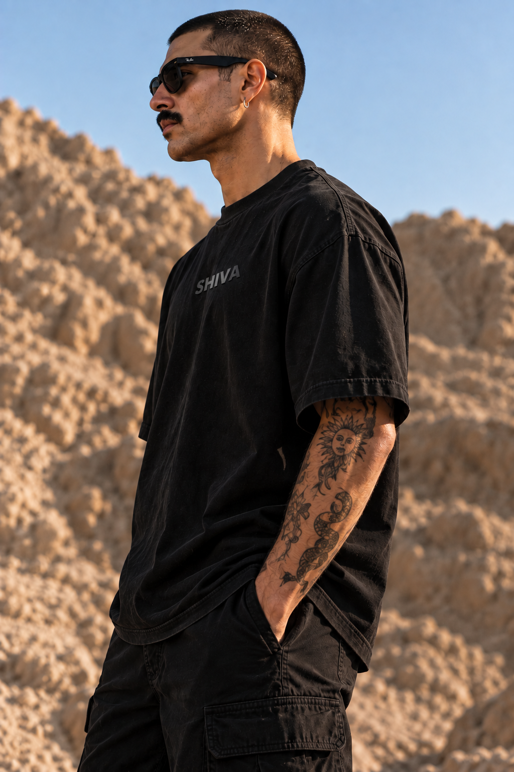 A man wearing a black oversized LMNO t-shirt with 'SHIVA' printed on the chest, standing outdoors against a rocky desert background.