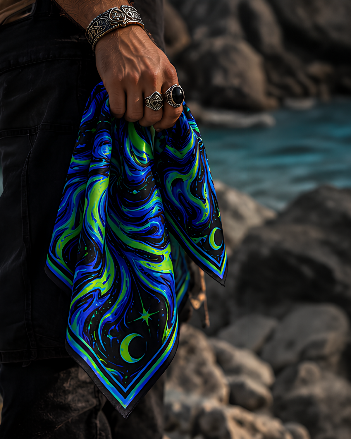 A person outdoors by the water holds a square LMNO scarf with a swirling cosmic print in blue, green, and black. The person is wearing silver rings and a silver bracelet.