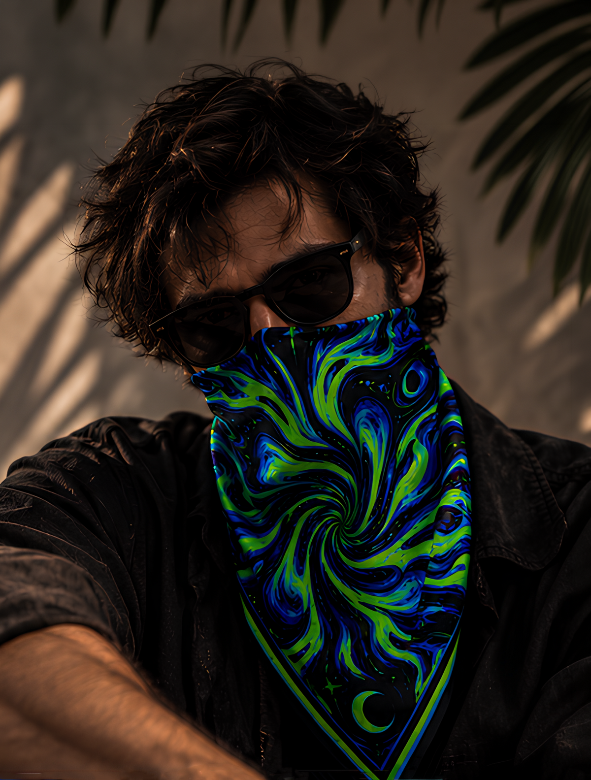 A man wearing a black shirt and sunglasses, with a vibrant blue and green marbled LMNO bandana covering his lower face, set against a warm-toned background with leaf shadows.