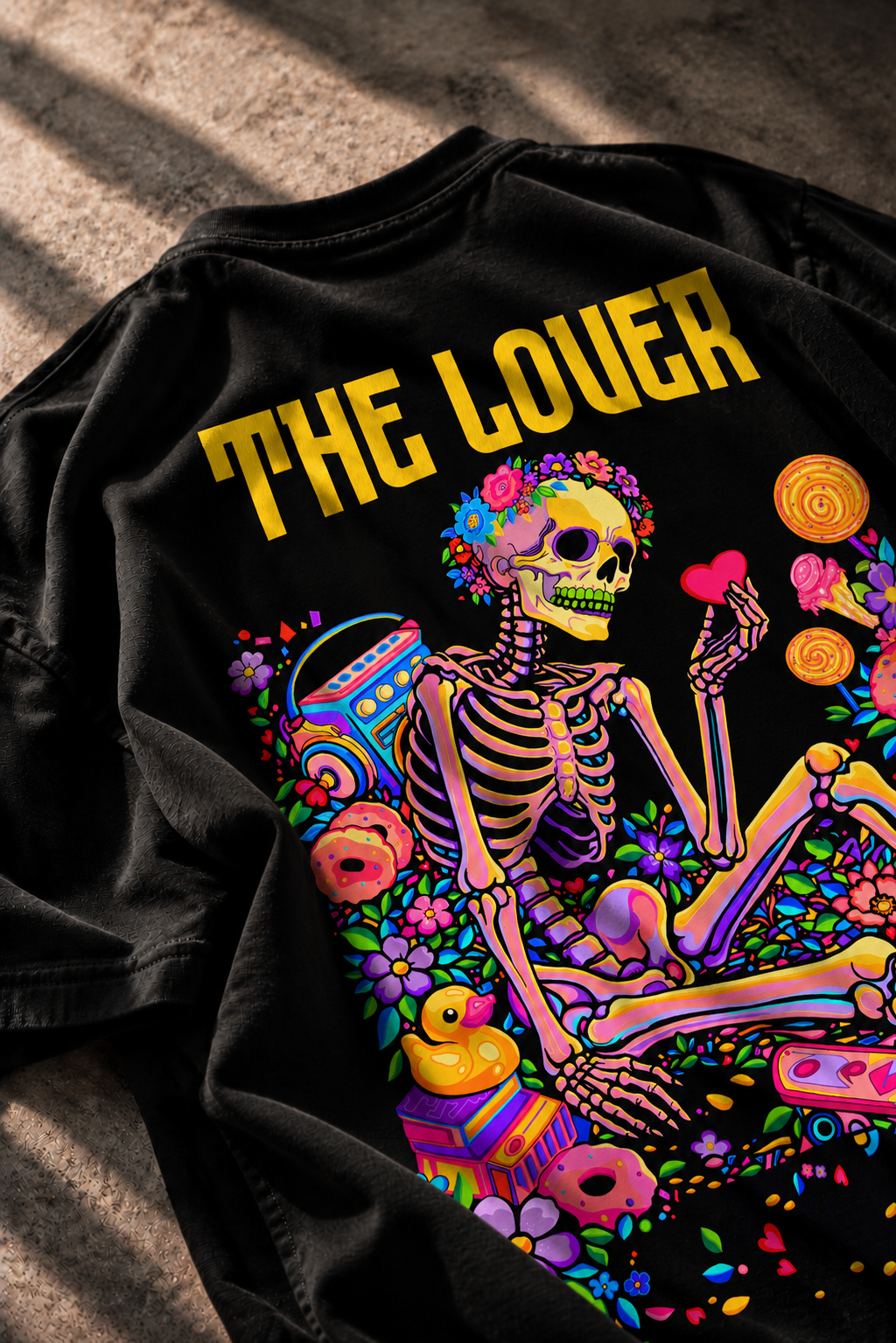 A black graphic LMNO t-shirt laid flat on a textured concrete surface. The shirt features a vibrant, colorful Day of the Dead style design of a skeleton surrounded by flowers, donuts, a boombox, and a yellow rubber duck, with the text 'THE LOVER' printed in yellow above the graphic.