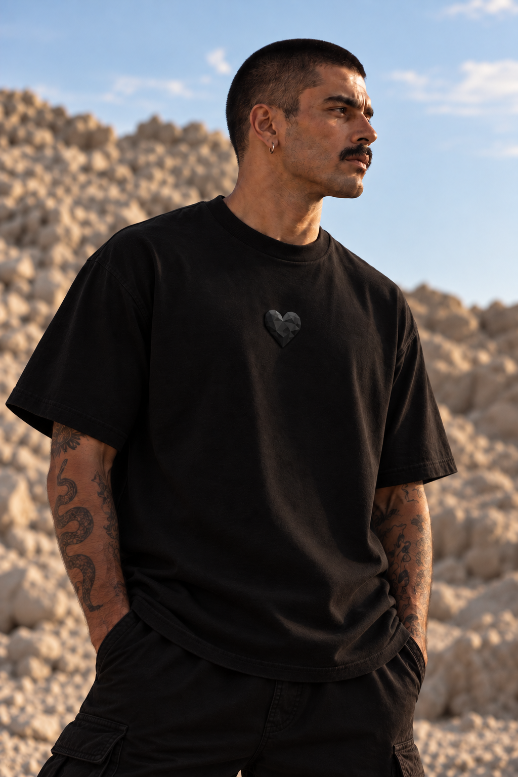 A man wearing a black LMNO t-shirt with a small black heart graphic on the left chest, standing outdoors against a rocky desert background under a blue sky.