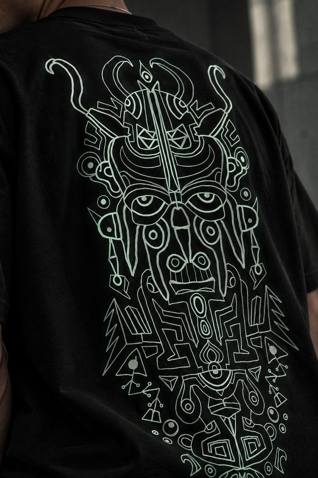 A close-up of a person wearing a black T-shirt, showing the back of the shirt with a large light green geometric tribal face graphic.