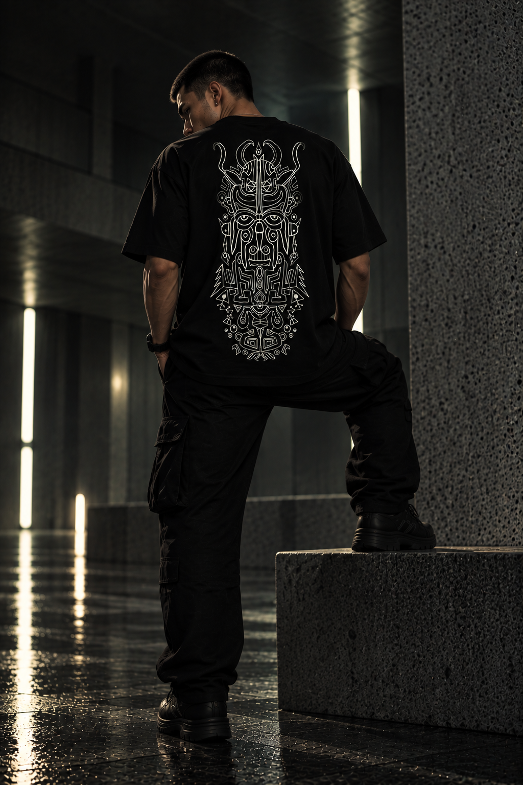 A man wearing a black LMNO t-shirt with a large white graphic print on the back, paired with black cargo pants, standing on a concrete ledge in a dark, moody urban setting.