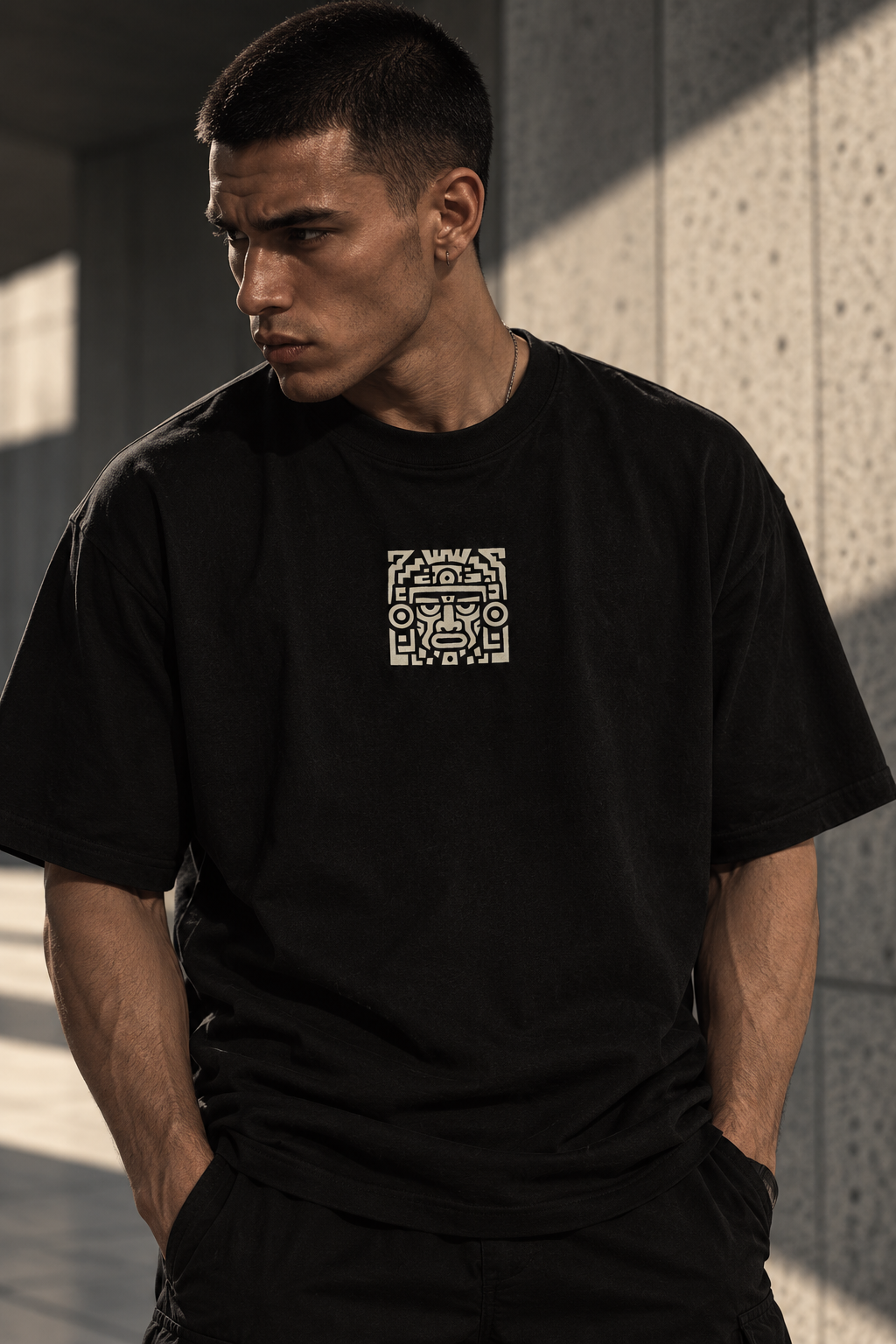 A man wearing a black LMNO t-shirt with a small white graphic print on the left chest, standing against a textured concrete wall with dramatic shadows.