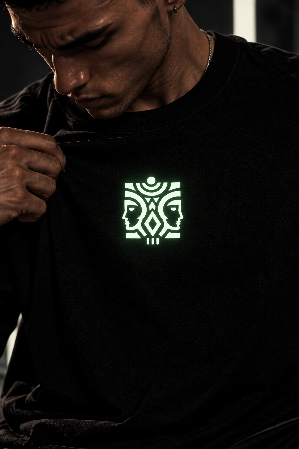 A close-up of a man wearing a black LMNO t-shirt, pulling the fabric to reveal a glowing green graphic on the chest featuring two stylized faces and geometric patterns.