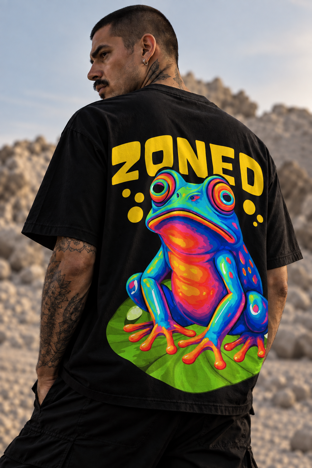 A man wearing a black LMNO t-shirt with a large, colorful graphic of a frog and the word 'ZONED' in yellow on the back, photographed outdoors against a rocky desert background.
